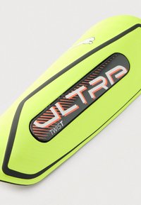 Φωτεινή κίτρινη ποδοσφαιρική προστατευτική επένδυση κνήμης με μαύρη ρίγα και κεντρική περιοχή λογότυπου που παρουσιάζει το "ULTRA TWIST" με λευκά και πορτοκαλί γράμματα.