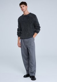 Maglione nero lavorato a maglia con una texture a costine, abbinato a pantaloni grigi chiari e scarpe nere. Il modello è in piedi contro uno sfondo neutro.