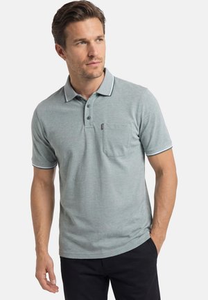 Mann trägt ein hellgraues Polohemd mit kurzen Ärmeln und Brusttasche sowie dunkle Hosen, schaut leicht nach links und hat die Hand in der Tasche.