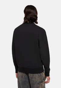 Personne aux cheveux ondulés et foncés portant un sweat-shirt noir à manches longues et un jean en denim bleu délavé, debout de dos devant un fond uni.
