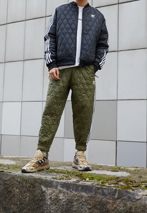 Personne portant une veste matelassée noire Adidas, un pantalon matelassé vert avec des bandes blanches et des baskets beige à motifs, debout à l'extérieur.