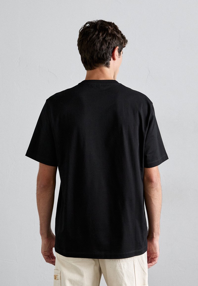 Man met donker haar die een eenvoudig zwart T-shirt met korte mouwen en lichtbeige cargobroek draagt, met zijn gezicht naar een grijze muur gekeerd.