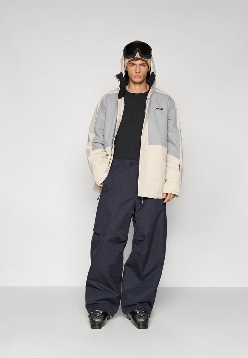 Ski jacket in beige and gray, with a hood. Paired with black long-sleeve top and navy cargo pants. Features snow goggles and ski boots.