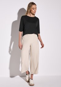 Černé tričko s krátkým rukávem v kombinaci s béžovými kalhotami s širokými nohavicemi a světlými teniskami. Oblečení má jednoduchý design a uvolněný střih.