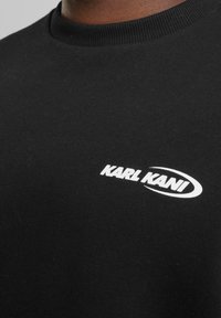 Black cotton sweatshirt with a round neck, featuring a white "KARL KANI" logo and smooth texture, highlighting simplicity in design.
