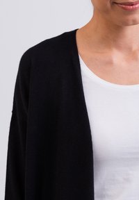 Svart cardigan i mjukt stickat material med öppen framsida, bärs över en vit T-shirt och visar en avslappnad passform samt en slät textur.