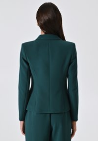 Blazer turchese con una vestibilità sartoriale, reverse e pince posteriori. Tessuto liscio con una leggera lucentezza. Indossato con pantaloni coordinati.