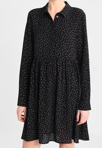 Robe noire à manches longues, col et taille froncée. Présente un motif à pois gris clair. Tissu léger et fluide.