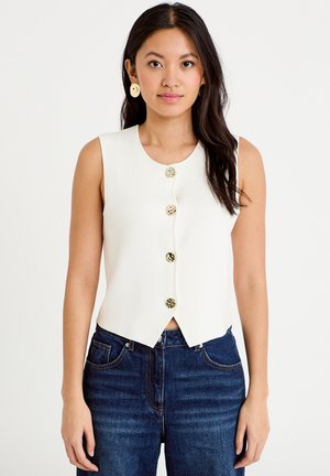 Młoda kobieta o długich ciemnych włosach, nosząca złote kolczyki w kształcie dysków, białą, bezrękawnikową bluzkę na guziki oraz ciemnoniebieskie dżinsy, zwrócona przodem.