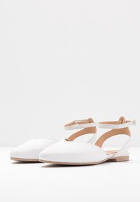 Białe płaskie buty z czubkiem w kształcie szpica, wyciętym wzorem, paskiem na kostkę i niskim obcasem, wykonane z gładkiej skóry i o minimalnych detalach.
