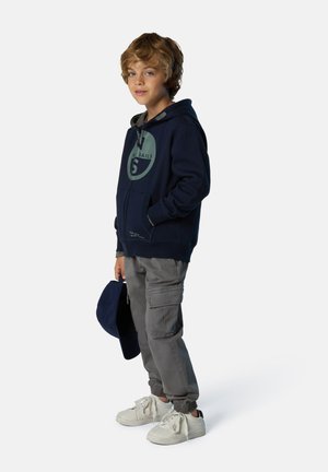 Felpa con cappuccio blu navy con un logo circolare, pantaloni cargo grigi eSneakers bianche; ragazzo che tiene in mano un cappellino blu abbinato.