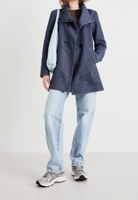 Marineblauer Wollmix-Doppelbreasted-Mantel mit Reverskragen, kombiniert mit hellblauen Jeans und grauen Sneakers. Eine hellblaue Tasche setzt einen Kontrast.
