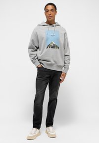 Grå hættetrøje med bjerggrafik, snøre i hætten og ribbede manchetter, parret med sorte jeans og hvide sneakers med brune detaljer.