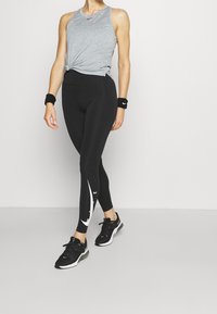 Sorte leggings med hvid Swoosh-grafik, kombineret med en grå ærmeløs top og sorte sneakers. Sort armbånd. Glat stoftekstur.