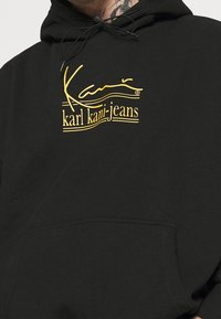 Felpa nera realizzata in misto cotone, con un logo stampato giallo che combina testo in corsivo e in stampatello. Presenta una tasca frontale e cordini regolabili.