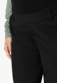 Pantalons noirs avec une texture lisse et une taille plate dotée d'une passante de ceinture, associés à un haut à manches longues vert à côtes.