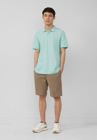 Polo shirt vert menthe avec un col, associé à un short beige et des baskets blanches. Le tissu semble lisse et respirant. Design minimaliste.