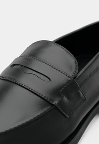 Gros plan d'un mocassin en cuir noir avec une lanière décorative et des coutures détaillées sur un fond clair uni.