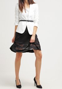 Valkoinen muotoiltu bleiseri syvällä V-puuvilla, yhdistetty mustiin hameeseen, jossa on pitsiset paneelit ja kerrostettu muotoilu. Mustat korkokengät täydentävät asua.