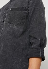 Camicia in denim nero con tasca sul petto, design con bottoni, maniche arrotolabili fissate con bottoni a pressione e una texture leggermente sbiadita.