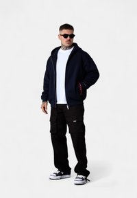 Haut à zip bleu marine avec deux poches latérales, T-shirt blanc, pantalon cargo noir et baskets noires. Présente un accent rouge contrastant dans les poches.