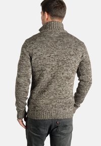Strikket genser i teksturerte brune og kremfarger, med høy krage og ribbestrikkede mansjetter og kant, kombinert med mørkegrå jeans.