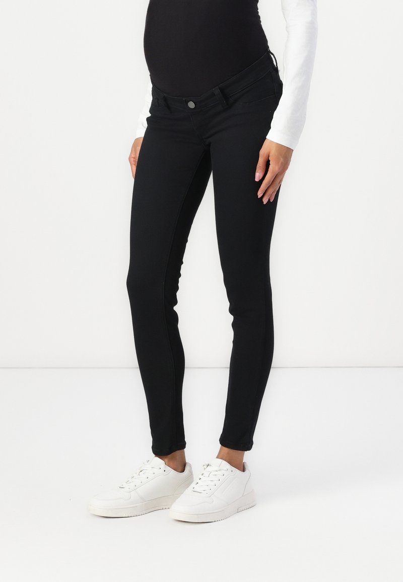 Personne enceinte portant un jean de maternité noir, un haut à manches longues blanc et des baskets blanches, se tenant devant un fond blanc uni.