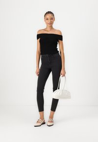Juodas off-the-shoulder ribotas viršus, tamsūs siauri džinsai, šviesios spalvos rankinė su dviem diržais ir smėlio spalvos butai su dirželių akcentais.