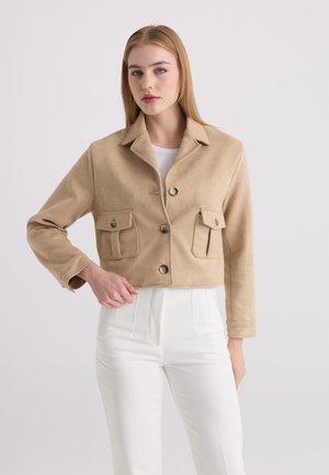 Giovane donna con capelli biondi lisci che indossa una giacca corta beige con bottoni e tasche con patta, abbinata a pantaloni bianchi a vita alta.