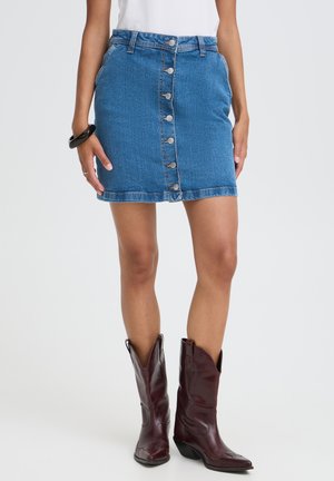 Kvinde iført midlang blå denim nederdel med knapper foran, hvid top, sort armbånd og mørkebrune læder cowboystøvler, stående foran en hvid baggrund.