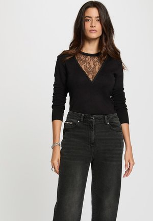 Maglione nero con inserto in pizzo al collo, maniche lunghe e polsini aderenti. Indossato con jeans in denim scuro a vita alta caratterizzati da una chiusura con bottone.