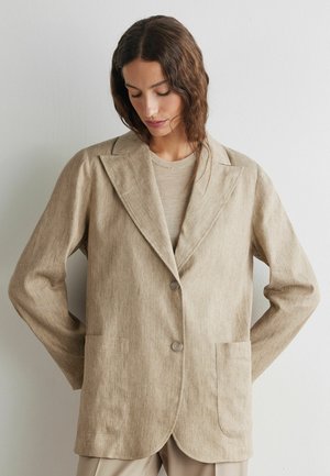 Kvinde iført oversize beige hørblazer med to knapper og store forlommer, kigger nedad mod en ensfarvet væg.