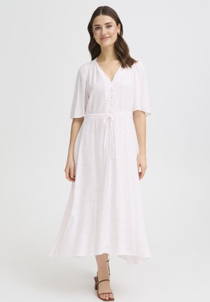 Femme portant une robe midi blanche à petits pois rouges, manches courtes, col en V, ceinture à la taille, et sandales à talons marron, debout sur un fond blanc.