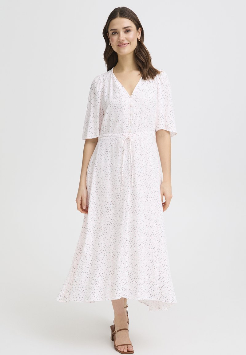 Femme portant une robe midi blanche à petits pois rouges, manches courtes, col en V, ceinture à la taille, et sandales à talons marron, debout sur un fond blanc.