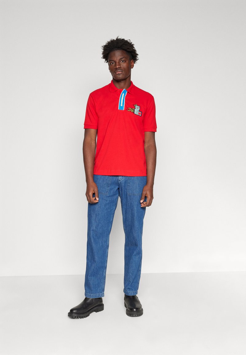 Polo rouge avec un col à accents bleus, présentant un design graphique. Porté avec un pantalon en denim bleu et des chaussures noires. Texture de tissu lisse.