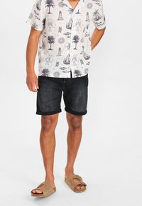 Košile s krátkým rukávem na bílém pozadí s námořnickými vzory zvířat a námořních prvků, kombinovaná s tmavými denimovými šortkami a hnědými sandály.