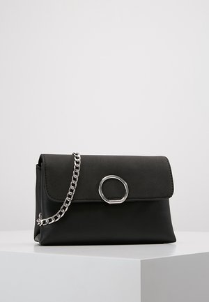 Zwarte leren clutch met klep, zilveren cirkelvormige sluiting en zilveren kettingriem, geplaatst op een wit voetstuk tegen een effen achtergrond.