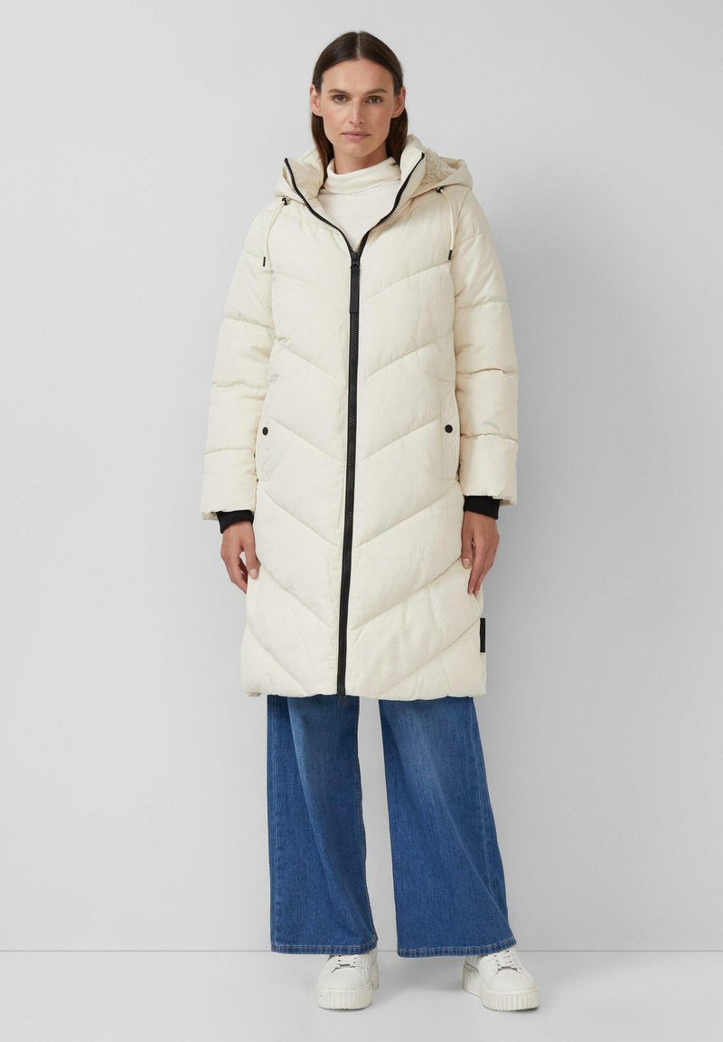 Pufīgs krēma garais mēteli ar zvīņainu rakstu, melns rāvējslēdzis, kapuce un sānu kabatas, kombinēts ar platu džinsu biksēm un baltām sporta apavām.