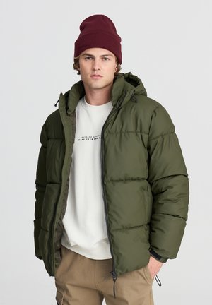 Giovane uomo che indossa un berretto bordeaux, una giacca a vento verde aperta sopra una felpa bianca e pantaloni beige con le mani in tasca.