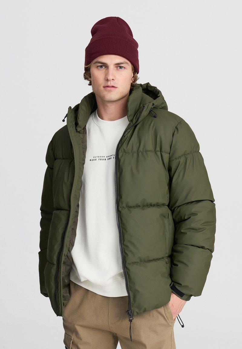 Giovane uomo che indossa un berretto bordeaux, una giacca a vento verde aperta sopra una felpa bianca e pantaloni beige con le mani in tasca.