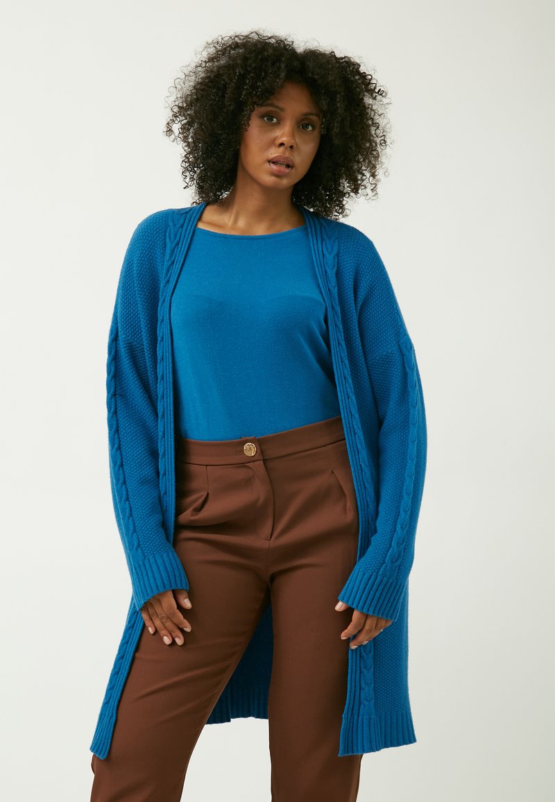 Cardigan lungo blu lavorato a maglia con dettagli a trecce, abbinato a una maglia blu e a pantaloni marroni a vita alta, caratterizzati da una texture liscia.