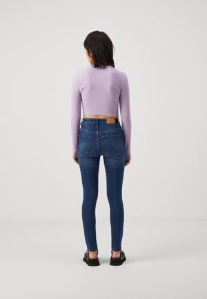 Mujer de espaldas con top corto de manga larga lila, vaqueros pitillo azul oscuro y sandalias negras sobre fondo blanco liso.