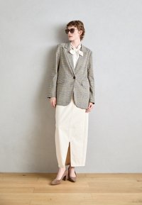 Beige és fekete kockás blézer, hosszú krémszínű szoknyával, amelynek elülső része hasított. A modellen barna magassarkú cipő és napszemüveg van.