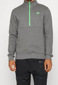 Šedá zipová mikina s vysokým límcem, která má jasně zelený zip a logo Nike. Vyrobena z měkkého materiálu a má standardní střih.