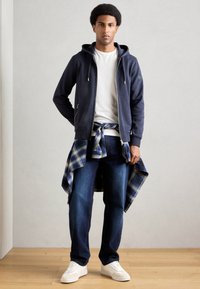 Mornarska zip-up hoodie, bijela majica, plavi karo flanel vezan oko struka, tamni traperice, bijele tenisice. Glatka tekstura, ležeran stil.