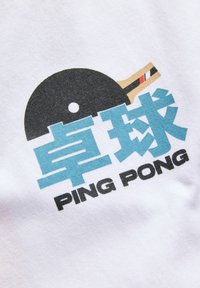 Biała koszulka z nadrukiem czarnej rakietki do ping-ponga oraz niebieskim tekstem w chińskich znakach i angielskim napisem „PING PONG”. 
