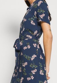 Woman wearing a navy blue floral dress with pink flowers, short sleeves, and a tied waist belt, showing a tattoo on her upper arm.