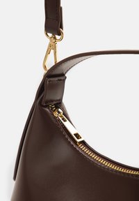 Σκούρα καφέ δερμάτινη τσάντα χειρός με χρυστό φερμουάρ και κούμπωμα στο λουράκι, εμφανίζεται σε κοντινό πλάνο πάνω σε απλό φόντο.