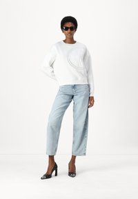 Sweatshirt bianco con logo in rilievo sul davanti, abbinato a jeans ampi azzurri e scarpe col tacco nere. Gli accessori includono occhiali da sole.