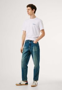 Hvid crew-neck t-shirt med blåt logo, lyseblå afslappede jeans og multicolore sneakers med hvide, sorte og røde accenter.